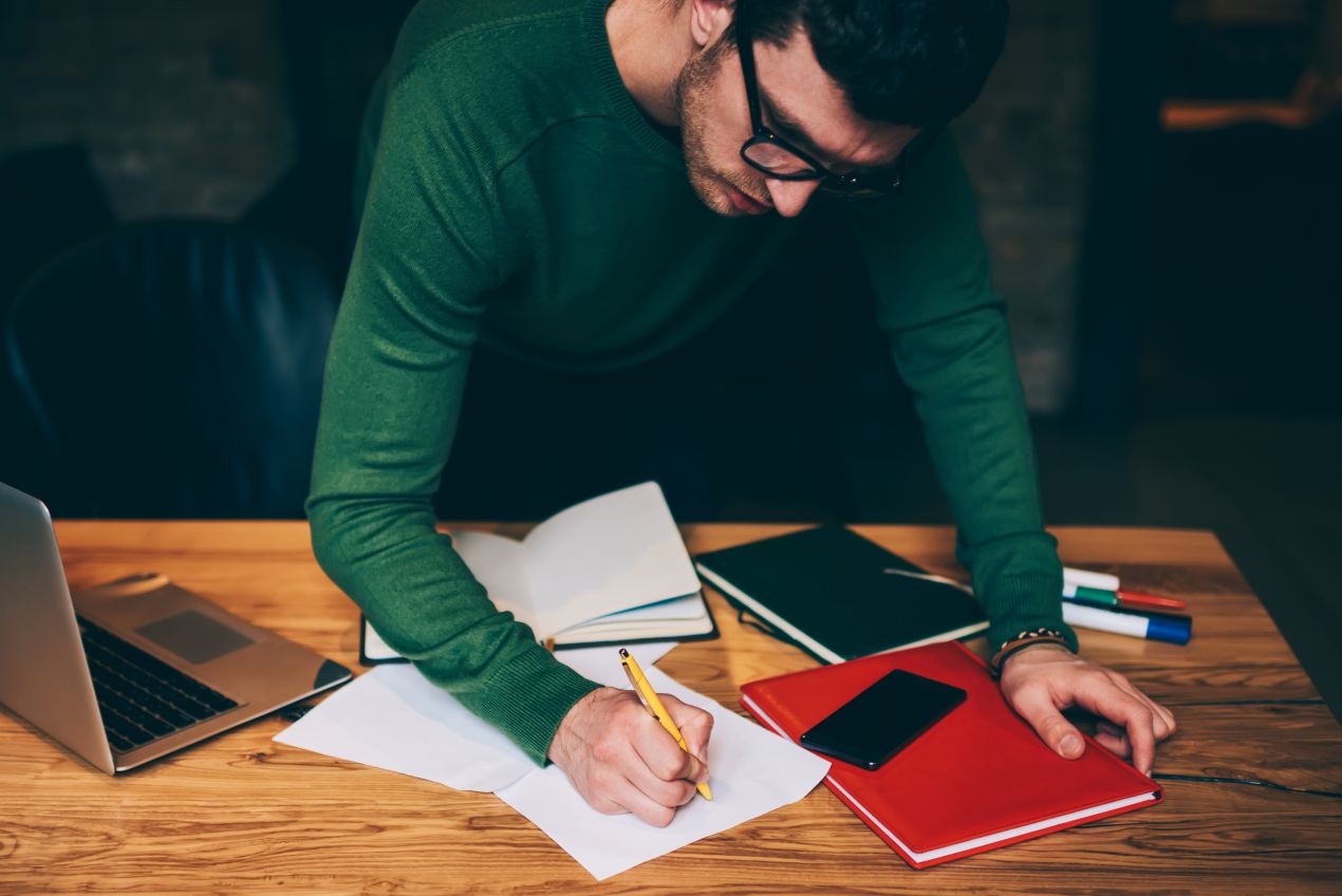 Ein Mann schreibt konzentriert an einem Tisch mit Laptop und Büchern. Ghostwriter Masterarbeit Erfahrungen zeigen, wie professionelle Hilfe abläuft.