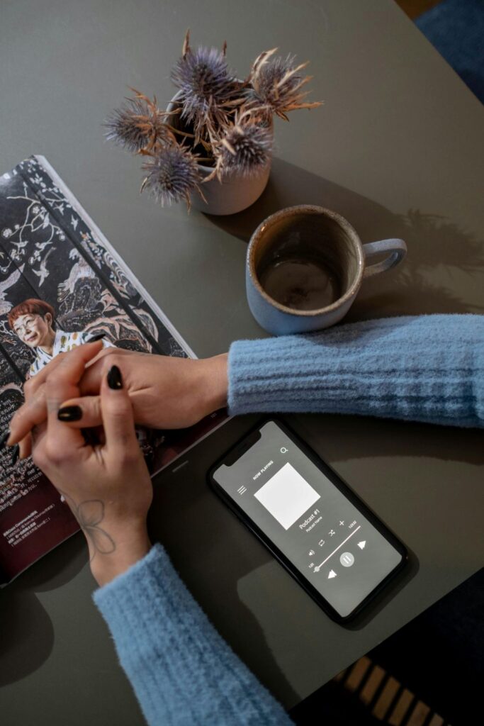 Person hoert KI-generierten Lernpodcast am Smartphone bei Kaffee, mit Zeitschrift und Trockenblumen auf Tisch
