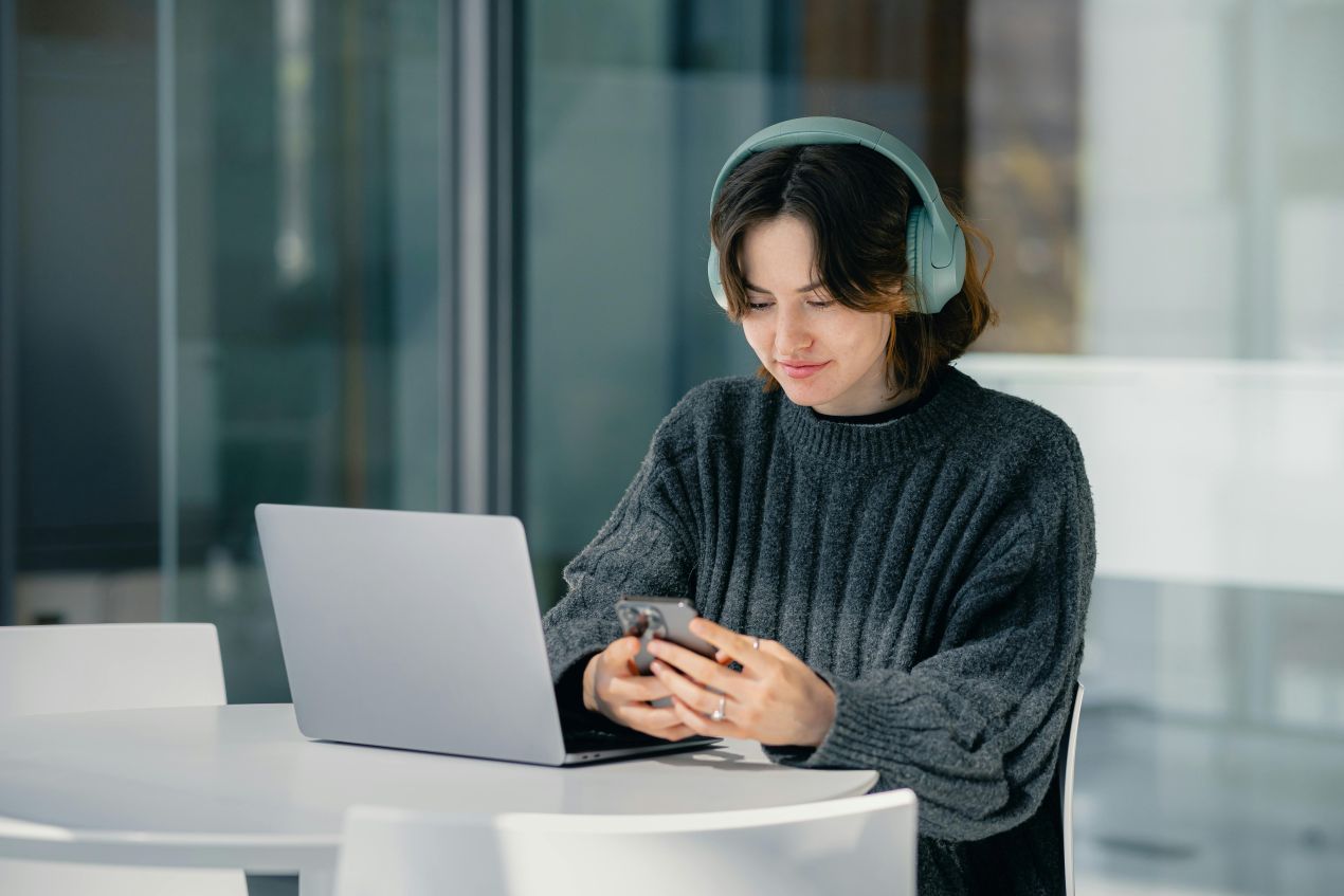 Junge Frau mit Laptop, Kopfhoerern und Smartphone erstellt Lernpodcast mit Google NotebookLM am Tisch in heller Umgebung