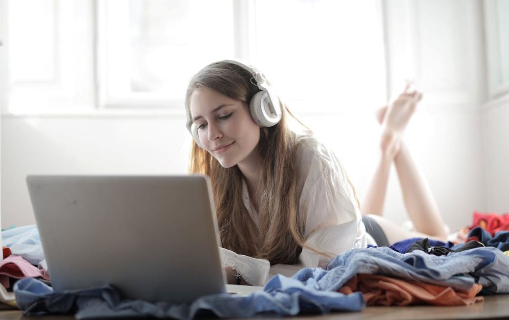 Junge Frau liegt entspannt mit großen Kopfhörern vor einem Laptop und hört ASMR in einem hellen Zimmer.